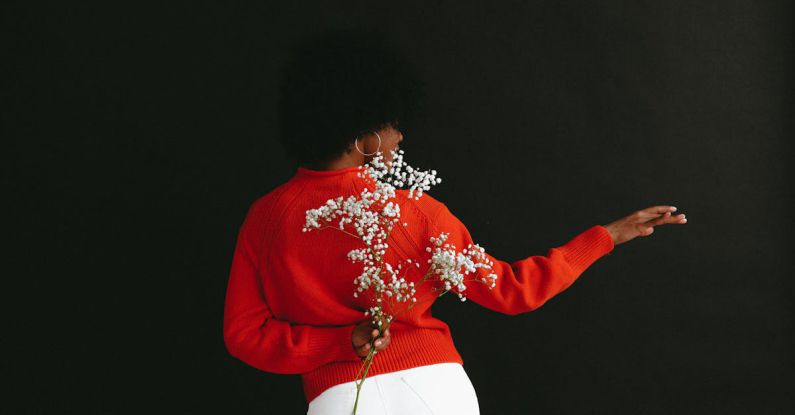 Music And Dance - Back view of sensual black woman in white denim and white red sweater holding Gypsophila flower behind back posing on black backdrop