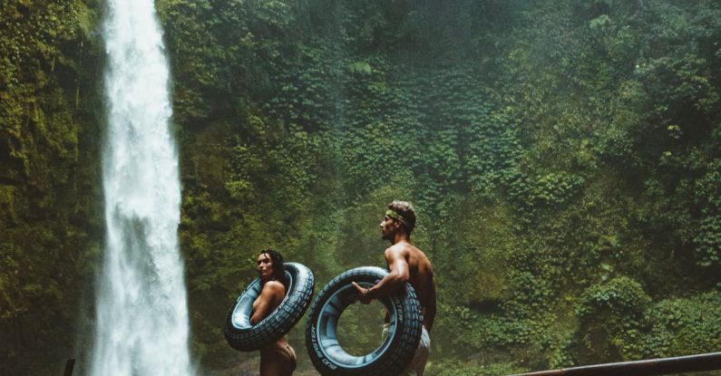 Adventures - Two Person Carrying Black Inflatable Pool Float on Brown Wooden Bridge Near Waterfalls